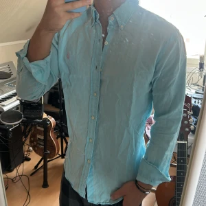 Ljusblå linneskjorta från Ralph Lauren - 100% Linen Snygg ljusblå skjorta från Ralph Lauren i linne med klassisk button-down krage och rosa broderad logga på bröstet. Skjortan har lång ärm och knäppning framtill. Perfekt för dig som gillar en fräsch och stilren look.