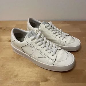 Vita sneakers från Golden Goose - Säljer ett par stilrena vita sneakers från Golden Goose, modell Stardan. Skorna är i skinn med klassisk stjärndetalj på sidan, snörning och diskreta loggor på häl och sula. Perfekta för dig som gillar minimalistisk och clean stil.
