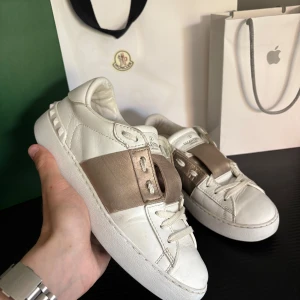 Valentino Garavani open sneakers - Snygga vita Valentino Garavani Rockstud sneakers i skinn med metallic guldbrun detalj över snörningen. Klassisk rund tå, platt sula och ikoniska nitar baktill. Perfekta för dig som vill ha en lyxig och trendig look.