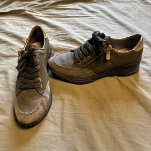 Grå sneakers från Rieker med dragkedja - Snygga grå sneakers från Rieker med detaljer i mocka och skinn. Skorna har svarta snören, guldfärgad dragkedja på sidan och en robust svart sula. Perfekta för dig som gillar en sportig och stilren look med extra komfort.