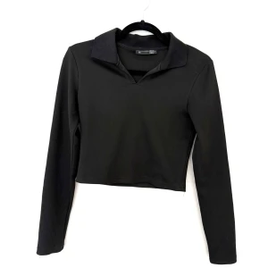 Svart ribbad croppad tröja Dazy - Svart croppad tröja från Dazy med lång ärm och klassisk krage. Tröjan är ribbad och har en stilren, enkel look som passar perfekt till jeans eller kjol. Materialet är mjukt och stretchigt för en bekväm passform.