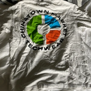 Vit Chinatown Market t-shirt med tryck - Vit t-shirt från Chinatown Market med stort färgglatt tryck på ryggen i grönt, blått och rött samt svart text. Liten logga framtill på bröstet. Klassisk passform och rund hals. Perfekt för dig som gillar streetwear vibes. Lite smutsiga prickar på några ställen om man kollar noga. 
