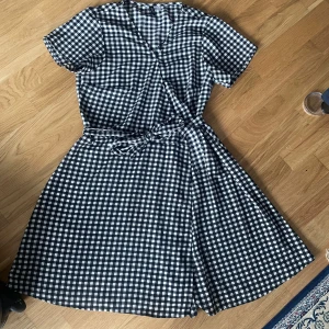 Rutig omlottklänning från H&M - Supersöt svartvit rutig kortklänning från H&M med omlottmodell och knytband i midjan. Klänningen har korta ärmar och v-ringning, perfekt för en chill sommardag. Tunt och skönt bomullstyg som känns luftigt och bekvämt. Passar 34-38 s-M