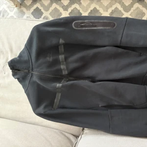 Svart Nike teach med huva samt byxor/ Dress - Svart dress från Nike med hel dragkedja, huva och diskreta svarta detaljer. Jackan har en ficka med dragkedja på ärmen och två svarta linjer över bröstet. Perfekt för dig som gillar sportig och clean stil. 