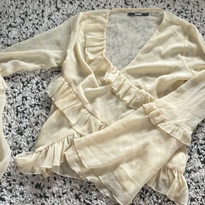 Beige omlottblus med volang från BikBok - Supersöt ljusgul blus från BikBok med volangdetaljer längs v-ringningen, ärmsluten och nederkanten. Tunn och luftig med lång ärm, perfekt för vår och sommar. Är i storlek s men passar även till xs.