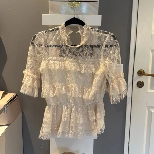 blus med volanger - Superfin ljus spetsblus med volanger och hög hals. Blusen är genombruten med vackra spetsdetaljer och har halvlånga ärmar med volangkant. Perfekt för dig som gillar en romantisk och feminin stil.