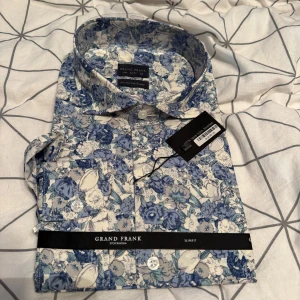 Blommig skjorta från Grand Frank - Snygg skjorta från Grand Frank med blått och vitt blommönster. Slim fit-modell i storlek L med klassisk krage och knappar framtill. Perfekt för dig som vill sticka ut med en färgglad och stilren look. Aldrig använd.