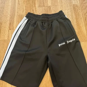 Palm Angels svarta shorts med ränder - Svarta shorts från Palm Angels med vita sidoränder och broderad logga på vänster ben. De har elastisk midja och sportig look, perfekta för en avslappnad streetstyle. Materialet känns mjukt och följsamt, och shortsen har fickor på båda sidor.