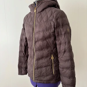 Brun dunjacka från Michael Kors S - Snygg brun dunjacka från Michael Kors med guldiga dragkedjor och huva. Jackan är quiltad och har ett diskret märke på ärmen. Yttermaterialet är nylon och fodret är polyester, fylld med dun. Perfekt för kyliga dagar när du vill vara både varm och stilren.