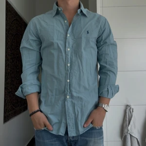 Ljusblå skjorta från Ralph Lauren - Klassisk ljusblå skjorta från Ralph Lauren med broderad logga på bröstet. Skjortan har lång ärm, knappar framtill och en avslappnad passform. Perfekt till jeans och enkel att styla för en clean look.
