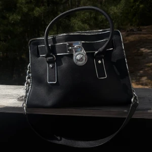 Svart handväska från Michael Kors - Stilren svart handväska från Michael Kors med silverfärgade detaljer och kedja på axelremmen. Väskan har ett klassiskt MK-lås framtill och både handtag och axelrem för flexibel användning. Perfekt för dig som gillar en tidlös och lyxig look.