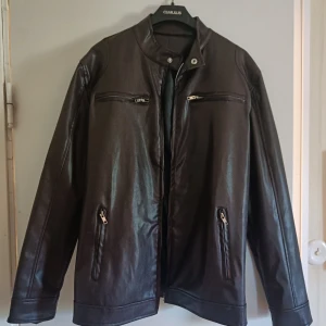 Brun skinnjacka i konstläder med dragkedjor - Snygg mörkbrun jacka i konstläder med stilren design och ståkrage. Ny (men prislapp ej kvar), ej använd pga liten storlek. Jackan har två fickor med dragkedjor och två dragkedjor för dekoration framtill samt rak passform. Perfekt för dig som vill ha en klassisk look med edge. Långärmad och passar till många olika outfits.