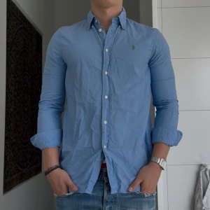 Blå skjorta från Ralph Lauren M - Klassisk ljusblå skjorta från Ralph Lauren i relaxed fit. Skjortan har långa ärmar, knappar framtill och ikonisk grön logga broderad på bröstet. Tillverkad i bomull och passar perfekt till jeans för en clean look.