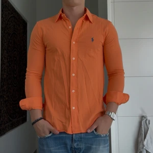 Orange skjorta från Ralph Lauren - Snygg orange skjorta från Ralph Lauren med klassisk krage och långa ärmar. Skjortan har knappar framtill och ikonisk blå broderad logga på bröstet. Tillverkad i mjuk bomull och har en slim passform som sitter riktigt bra. Perfekt nu till sommarn. Passar både med en vit tisha under eller bara skjortan i sig! Storlek S men kan även passa M (jag är M)