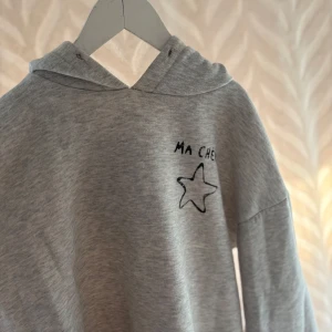 Grå hoodie med tryck 'Ma Chérie' - Säljer en ljusgrå hoodie med svart tryck där det står 'Ma Chérie' och en stjärna på både fram- och baksidan. Tröjan har huva och är tillverkad i mjuk bomullsmix. Perfekt för en chill och avslappnad stil.