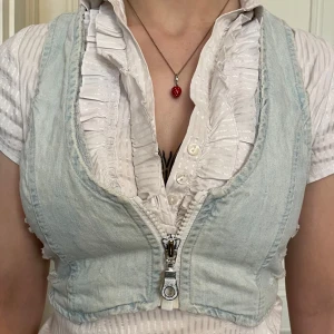 Ljusblå croppad jeansväst med dragkedja - Säljer en ljusblå croppad jeansväst med råa kanter och slitna detaljer på sidorna. Västen har en markerad dragkedja framtill och är ärmlös med djupa ärmhål. Perfekt för att ge din outfit en edgy touch.