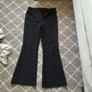 Svarta bootcut kostymbyxor RYVLS 158 - Svarta kostymbyxor från RYVLS i storlek 158. Byxorna har bootcut-modell med utsvängda ben och klassisk midja. Gjorda i ett mjukt och följsamt material, perfekta för en clean och stilren look. Justerbar midja med knapp på insidan.