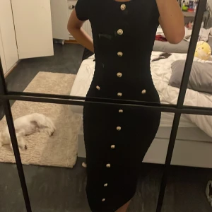 Svart långklänning med guldknappar - Supersnygg svart långklänning med tight passform och korta ärmar. Klänningen har dekorativa guldknappar framtill och en lätt ribbad struktur i tyget. Perfekt för dig som gillar en stilren och elegant look med en twist.