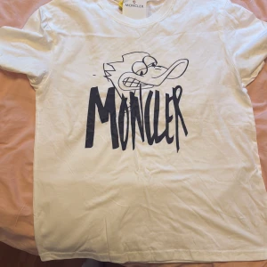 Vit Moncler t-shirt med tryck - Vit t-shirt från Moncler med coolt svart tryck framtill som föreställer en tecknad figur och stor Moncler-logga. Klassisk rund halsringning och korta ärmar. Skön och mjuk bomull som passar perfekt till jeans eller shorts. Tröjan är inte original, men bästa kopian på marknaden, den är i storlek M, men passar även S. Kontakta mig för mer information!