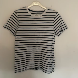 Randig t-shirt från H&M - Vit och svart randig t-shirt från H&M med klassisk rund halsringning. T-shirten har korta ärmar och är tillverkad i mjuk bomull, perfekt för en chill och stilren look.