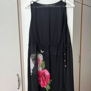 Svart blommig klänning med snörning - Svart ärmlös klänning med stora blommor i rosa och vitt nertill. Klänningen har smala axelband, snörning i midjan och ett lätt, luftigt tyg som ger en flowy känsla. Perfekt för dig som gillar romantiska detaljer och vill sticka ut med blommigt mönster.