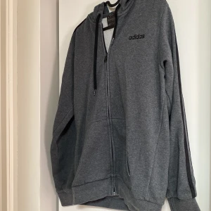 Grå zip-hoodie från Adidas - Tja folket! Jag säljer en schysst Mörkgrå hoodie från Adidas med dragkedja, huva med snörning och svarta detaljer längs ärmarna. Klassisk Adidas-logga på bröstet och två stora fickor framtill. Perfekt för chill eller träning. Den har små tecken på användning ungefär 3-5 gånger, skriv till mig ifall ni vore intresserade! 