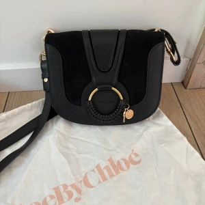 Svart axelväska från See By Chloé - Snygg svart axelväska från See By Chloé med guldfärgade metalldetaljer och rund ring framtill. Väskan är tillverkad i en mix av skinn och mocka, har justerbar axelrem och magnetlås. Perfekt storlek för det viktigaste och stilren design med exklusiv känsla. Mått: 21.5*19*5.5cm