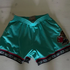 Turkosa Memphis Grizzlies shorts Mitchell & Ness - Snygga turkosa NBA Hardwood Classics shorts från Mitchell & Ness med elastisk midja och ikoniskt Vancouver Grizzlies-björntryck på benet. Shortsen har svart, röd och vit mönstrad kant nertill och är gjorda i luftig mesh av polyester. Perfekta för basket eller streetwear.
