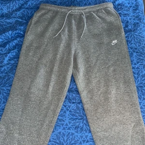 Grå mjukisbyxor från Nike, storlek M - Gråa mjukisbyxor från Nike i storlek M. Byxorna har dragsko i midjan, en bakficka och Nike-logga på vänster lår. Tillverkade i mjukt bomullsmaterial, perfekta för chill eller träning. Klassisk och enkel design med relaxed fit.