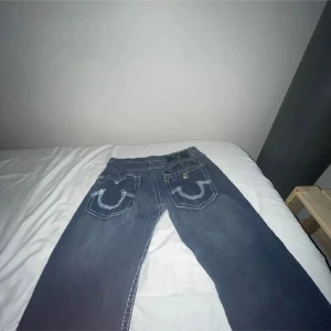 True Religion jeans med broderade fickor - Säljer ett par mörkblå jeans från True Religion med ikoniska broderade bakfickor och kontrastsömmar. Klassisk femficksmodell med raka ben och låg midja. Jeansen har en snygg tvätt och detaljerade sömmar som sticker ut. Skriv vid frågor! Prutning är löst genom text om intresse!