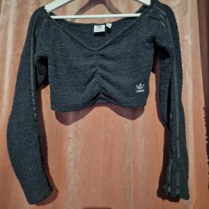 Svart croppad topp från Adidas - Svart croppad topp från Adidas i mjukt, frottéliknande material. Långärmad med v-ringning och snygg rynkdetalj framtill. Klassiska Adidas-loggan broderad på bröstet och tre diskreta ränder längs ärmarna. Perfekt för en sportig och trendig look. Ny utan prislapp. 
