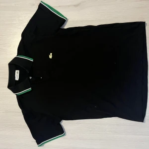 Svart Lacoste piké  - Tjena säljer  en svart Lacoste piké med gröna detaljer och snygg design som är perfekt till sommaren 