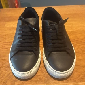 Svarta sneakers från Axel Arigato - Stilrena svarta sneakers från Axel Arigato med vit platt sula och svart snörning. Ovandel i skinn och detaljer i mocka vid hälen. Diskret guldtext på sidan och Arigato-logga bak på sulan. Perfekta för en clean och modern look.