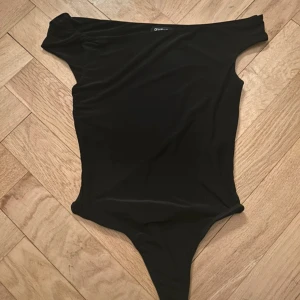 Svart one shoulder body - Säljer en svart body från Boohoo med one shoulder-design och twistad detalj vid axeln. Tight passform och tillverkad i stretchigt syntetmaterial som sitter snyggt på kroppen. Perfekt för dig som vill ha en stilren och trendig topp. Aldrig använd!