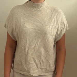 Beige stickad polotröja utan ärmar - Beige stickad polotröja med ribbad hög krage och ärmlös design. Tröjan har en loose fit och är perfekt för lager-på-lager. Mjuk och skön känsla, passar till både jeans och kjol. Enkel och stilren look som funkar året runt.