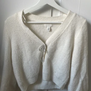 Vit stickad kofta med rosettdetalj - Superfin vit stickad kofta från H&M med croppad passform och v-ringning. Koftan har en snygg rosettdetalj framtill i silver och långa ärmar. Perfekt att slänga över en topp för en chill men ändå söt look.