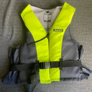 Gul och grå flytväst från Baltic - Säljer en flytväst från Baltic i grått och neongult med svart spännbälte och dragkedja framtill. Västen har en klassisk, sportig look och är perfekt för vattenaktiviteter. Märket syns tydligt på bröstet och insidan är ljusgrå.