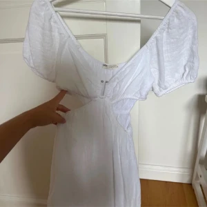Vit klänning  - Superfin vit kortklänning från pull and bear med v-ringning, puffiga kortärmar och cut-out detaljer vid midjan. Klänningen har en lätt och luftig känsla, perfekt för varma dagar. Tillverkad i bomull för extra komfort och sommarkänsla.