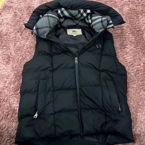 Svart dunväst från Burberry London - Snygg svart dunväst från Burberry London med klassiskt rutigt foder i huvan och dragkedja framtill. Västen har 3 fickor med dragkedja och en diskret Burberry-logga på bröstet. Perfekt för lager-på-lager-stil.