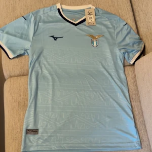 Ljusblå Lazio fotbollströja Mizuno - Snygg ljusblå Lazio fotbollströja från Mizuno med klubbmärke på bröstet och mörkblå detaljer vid krage och ärmslut. Tröjan har V-ringning och diskret mönster med klubbnamn i tyget. Perfekt för dig som älskar italiensk fotboll.