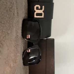  ÄKTA Svarta DG solglasögon från Dolce & Gabbana - Snygga svarta solglasögon från Dolce & Gabbana med stora, fyrkantiga bågar och DG-logga i silver med glittrande stenar på sidorna. Kommer med originalfodral. Perfekta för dig som vill sticka ut med en lyxig vibe.