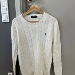 Vit kabelstickad tröja Polo Ralph Lauren - Vit kabelstickad tröja från Polo Ralph Lauren med klassisk rund halsringning och blå broderad logga på bröstet. Tröjan har lång ärm och ribbade muddar. Perfekt för dig som gillar en clean och tidlös stil.