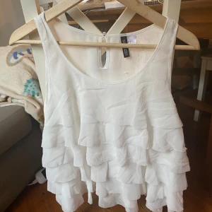Vit volangtop från H&M, strl 36 - Supersöt vit linnetopp från H&M med flera lager volanger som ger en flowy och luftig känsla. Toppen är ärmlös och har en rund halsringning samt en liten knappdetalj i nacken. Perfekt för sommarens alla tillfällen!