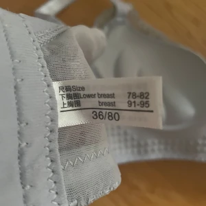 Grå spetsig bralette i storlek 36/80 - Säljer en snygg grå bralette med spets och blommigt mönster. Den har vadderade kupor, justerbara axelband och fina spetsdetaljer över hela toppen. Perfekt för dig som gillar en feminin och bekväm stil.