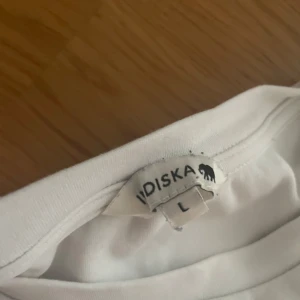 Vit t-shirt från Indiska, storlek L - Enkel och fräsch vit t-shirt från Indiska i storlek L. Klassisk rund halsringning och korta ärmar. Perfekt basic-plagg i mjuk bomull som funkar till allt. Snygg att styla med jeans eller kjol för en clean look.