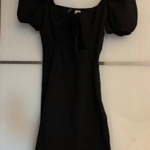 Svart kort klänning med puffärm och cut-out - Supersnygg svart kort klänning med puffärmar, knytning framtill och trendig cut-out under bysten. Klänningen har en figurnära passform och är tillverkad i ett mjukt material. Lite uppriven söm i armhålan på övre delen av dragkedjan.