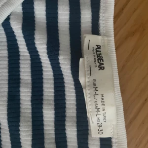 Randig ribbad linnetopp Pull&Bear - Säljer en ribbad linnetopp från Pull&Bear i storlek L. Toppen är vit med marinblå ränder och har en ärmlös design med breda axelband. Perfekt för sommardagar och enkel att matcha med jeans eller shorts.