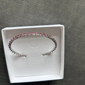 Caroline Svedbom armband  - Ett tunt och stilrent armband i silverfärg med små rosa stenar längs hela bandet. Armbandet är öppet i ändarna och har en minimalistisk design som ger en snygg och modern look. Perfekt för dig som gillar att addera lite färg till din outfit.