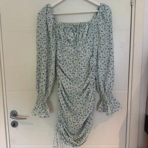 Adoore Capri klänning  - Supersöt ljusblå klänning från Adoore, fyrkantig ringning och långa volangärmar. Klänningen har snörning framtill och rynkade detaljer längs sidorna för en snygg passform. Perfekt för sommarens alla tillfällen! Endast använd 2 ggr och finns inte att köpa på Adoore längre.