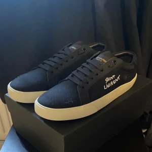 Svarta Saint Laurent sneakers - Svarta sneakers från Saint Laurent med vit sula och broderad logga på sidan. Klassisk låg modell med snörning och diskreta gulddetaljer på plösen. Ovandel i canvas och sula i gummi för en clean och stilren look.